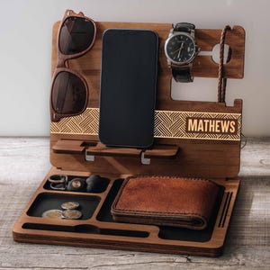 Könnte beinhalten: Ein hölzerner Schreibtisch-Organizer mit Fächern für Telefon, Uhr, Sonnenbrille, Geldbörse, Schlüssel und Münzen. Der Organizer trägt den beleuchteten Namen "MATHEWS" auf der Vorderseite. Die Sonnenbrille ist braun und die Geldbörse ist aus Leder.