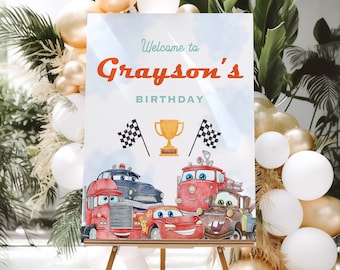 May include: A blue and white birthday sign with the text "Welcome to Grayson's Birthday" and a cartoon image of race cars. The sign is on a wooden easel in front of a backdrop of white and gold balloons.