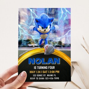 May include: A birthday invitation featuring the character Sonic the Hedgehog. The invitation has a blue and gold design with the name "NOLAN" in large blue letters. The text reads "IS TURNING FOUR" and includes the date, time, and location of the party.