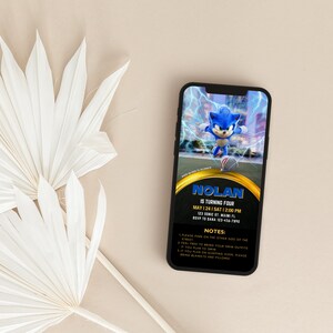 May include: A smartphone displays a birthday party invitation featuring Sonic the Hedgehog. The invitation has a black background with gold accents and includes the name "Nolan", party details, and notes. White dried palm leaves are in the foreground.
