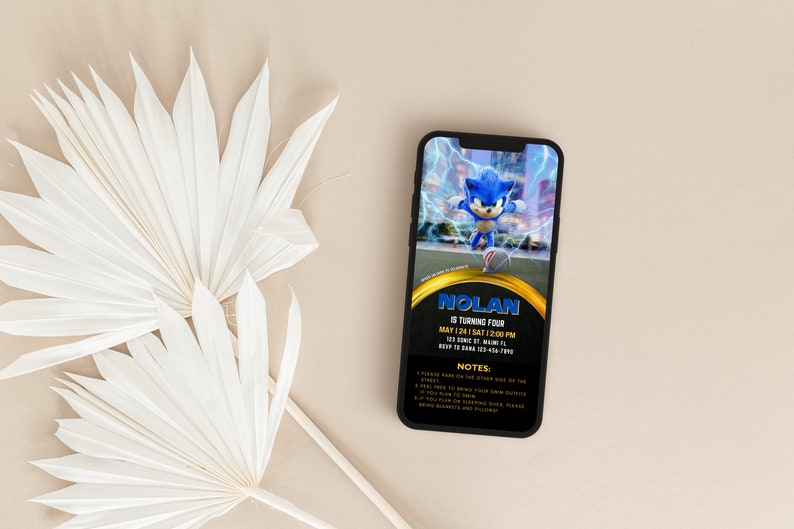 May include: A smartphone displays a birthday party invitation featuring Sonic the Hedgehog. The invitation has a black background with gold accents and includes the name "Nolan", party details, and notes. White dried palm leaves are in the foreground.