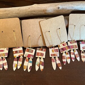 May include: Four pairs of dangle earrings with a geometric design. The earrings are made of white clay with red, orange, yellow, and blue stripes. The earrings are hanging from gold hoops.