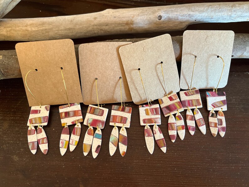 May include: Four pairs of dangle earrings with a geometric design. The earrings are made of white clay with red, orange, yellow, and blue stripes. The earrings are hanging from gold hoops.