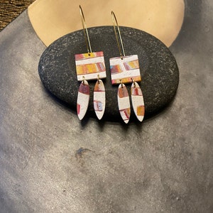 May include: A pair of dangle earrings with a geometric pattern in shades of red, orange, yellow, and white. The earrings are made of clay and have a gold-colored wire.