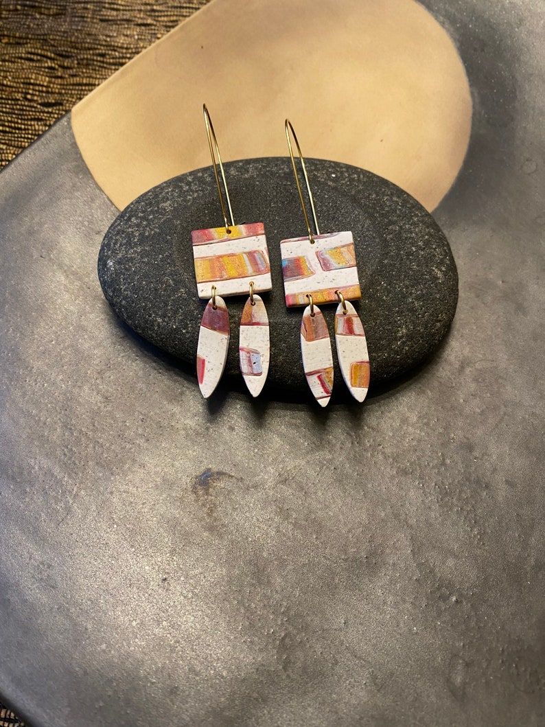May include: A pair of dangle earrings with a geometric pattern in shades of red, orange, yellow, and white. The earrings are made of clay and have a gold-colored wire.