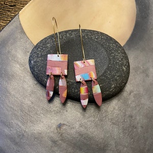 May include: A pair of dangle earrings with a geometric square design. The earrings are made of polymer clay and have a pink, white, and blue color scheme. The earrings are attached to gold-colored hooks.