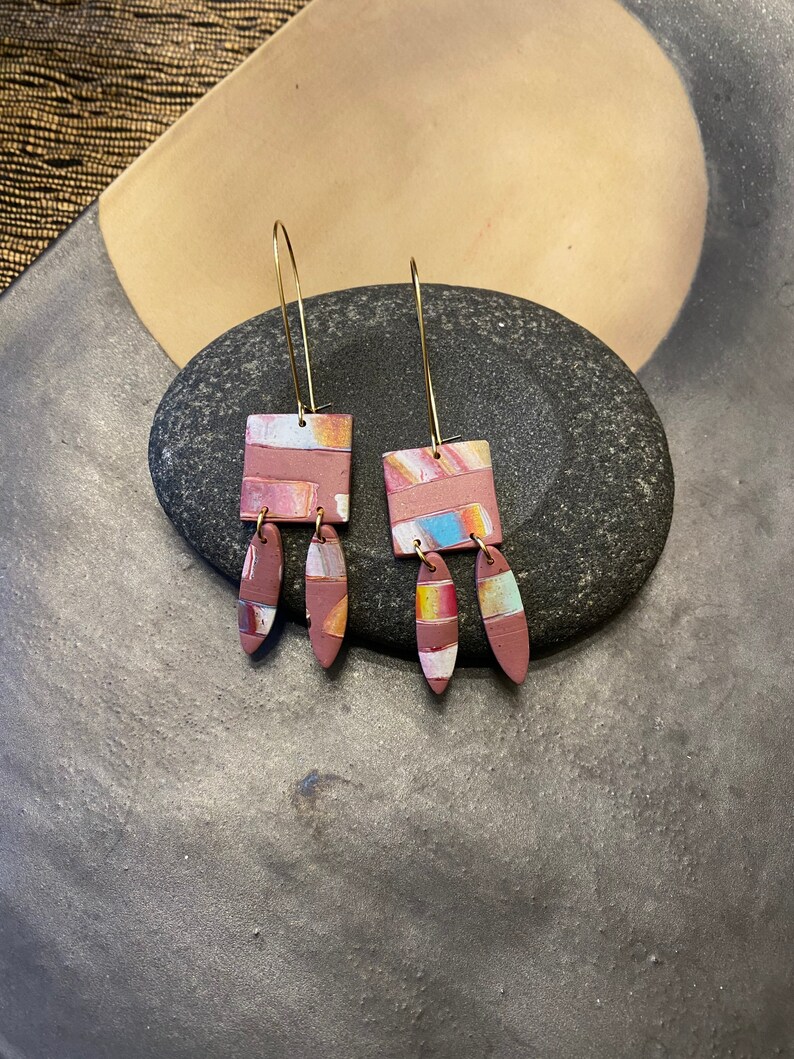May include: A pair of dangle earrings with a geometric square design. The earrings are made of polymer clay and have a pink, white, and blue color scheme. The earrings are attached to gold-colored hooks.