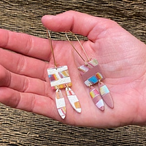 May include: A pair of dangle earrings with geometric shapes. The earrings are made of clay and have a white, pink, and brown color scheme. The earrings are attached to gold hoops.
