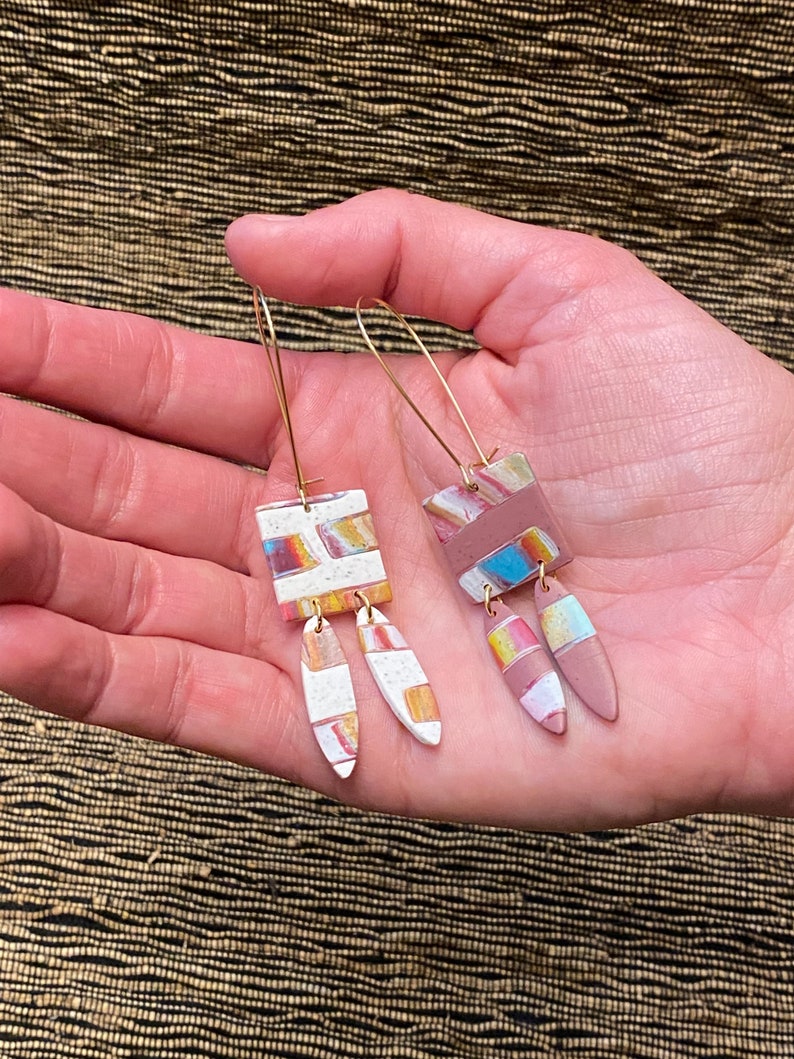 May include: A pair of dangle earrings with geometric shapes. The earrings are made of clay and have a white, pink, and brown color scheme. The earrings are attached to gold hoops.