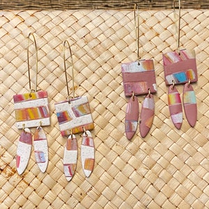 May include: Three pairs of dangle earrings with geometric shapes and abstract patterns in shades of pink, orange, and white. The earrings are made of polymer clay and have gold-toned hooks.