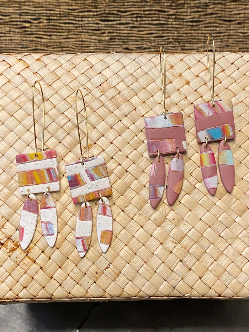 May include: Three pairs of dangle earrings with geometric shapes and abstract patterns in shades of pink, orange, and white. The earrings are made of polymer clay and have gold-toned hooks.
