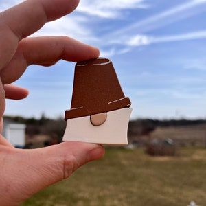 May include: A brown and white wooden gnome ornament with a brown hat and a white beard. The gnome is being held by a hand.