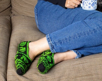 May include: A person wearing blue jeans and a black sweater sits on a couch with a mug in their hand. They are wearing green and black patterned socks.