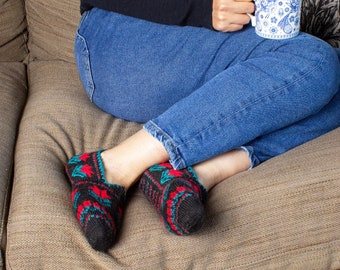 May include: A person wearing blue jeans and a black sweater sits on a sofa with a mug in their hand. They are wearing red and green knitted slippers.