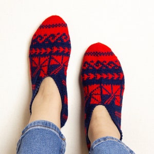 Könnte beinhalten: Ein Paar rote und blaue gestrickte Hausschuhe mit geometrischem Muster. Die Socken werden an den Füßen einer Person getragen, wobei die blauen Jeans der Person sichtbar sind.