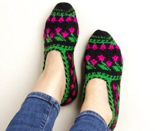 May include: A pair of black knit slippers with a floral pattern in pink and green. The slippers are worn by a person wearing blue jeans. The slippers have a green trim around the opening and along the sole.