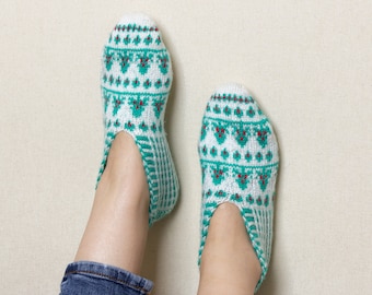 May include: A pair of white and green knitted slipper socks with a red and green pattern. The socks are being worn on a person's feet.