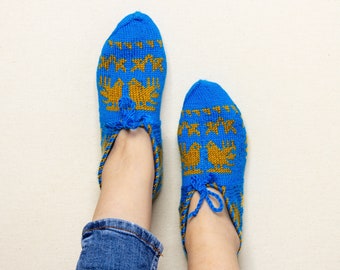 May include: A pair of blue knitted slippers with a gold bird and geometric pattern. The slippers have a tassel detail at the top and are worn with blue jeans. The slippers are a cozy and comfortable footwear option.