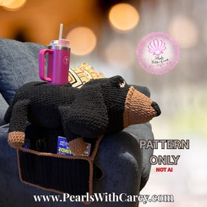 May include: A black and brown crochet dachshund armrest organizer with pockets, holding a pink tumbler with a straw. The organizer is on a gray couch arm. Text on the image reads "PATTERN ONLY NOT AI" and "www.PearlsWithCarey.com".
