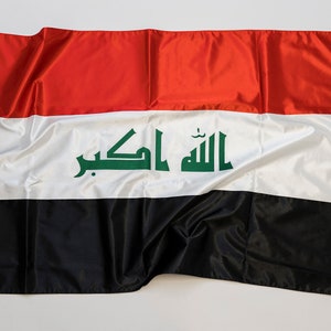 Op de afbeelding: Een rood, wit en zwarte vlag van Irak met de Arabische tekst "Allahu Akbar" in groen op de witte streep.