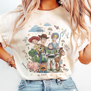May include: A cream-colored t-shirt featuring a colorful graphic print of Toy Story characters including Woody, Buzz Lightyear, Jessie, Rex, Hamm, Mr. Potato Head, Bo Peep, Bullseye, and Forky, with their names written in cursive script.