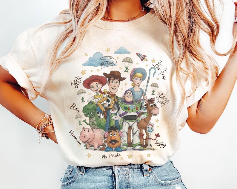 May include: A cream-colored t-shirt featuring a colorful graphic print of Toy Story characters including Woody, Buzz Lightyear, Jessie, Rex, Hamm, Mr. Potato Head, Bo Peep, Bullseye, and Forky, with their names written in cursive script.