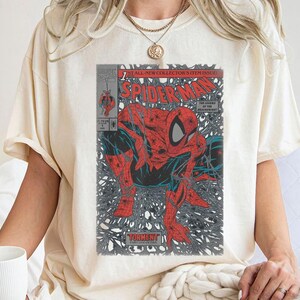 Pode incluir: Uma t-shirt de manga curta na cor creme com uma estampa gr&aacute;fica da capa de uma banda desenhada vintage do Spider-Man. A estampa mostra o super-her&oacute;i vermelho e azul numa pose agachada no meio de um padr&atilde;o de teia preta com o t&iacute;tulo Spider-Man.