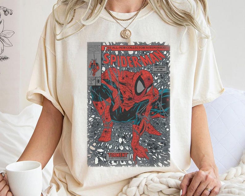 Pode incluir: Uma t-shirt de manga curta na cor creme com uma estampa gr&aacute;fica da capa de uma banda desenhada vintage do Spider-Man. A estampa mostra o super-her&oacute;i vermelho e azul numa pose agachada no meio de um padr&atilde;o de teia preta com o t&iacute;tulo Spider-Man.