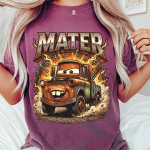 May include: A purple t-shirt featuring a graphic of Mater from the movie Cars. The design includes the word "MATER" in a distressed, metallic font above an illustration of the character. Lightning bolts and tools surround the image.