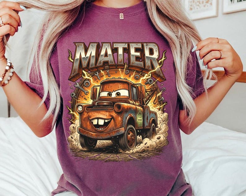 May include: A purple t-shirt featuring a graphic of Mater from the movie Cars. The design includes the word "MATER" in a distressed, metallic font above an illustration of the character. Lightning bolts and tools surround the image.