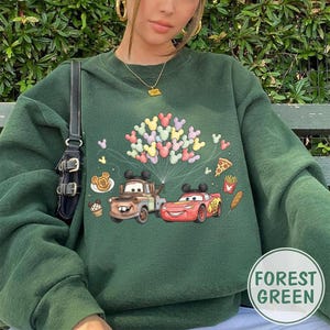 May include: Forest green sweatshirt with a graphic of Mater and Lightning McQueen from the Cars film, plus Mickey Mouse balloons and food icons. The sweatshirt has a crew neck and long sleeves.