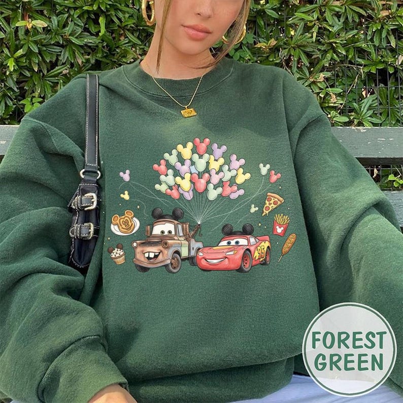 May include: Forest green sweatshirt with a graphic of Mater and Lightning McQueen from the Cars film, plus Mickey Mouse balloons and food icons. The sweatshirt has a crew neck and long sleeves.
