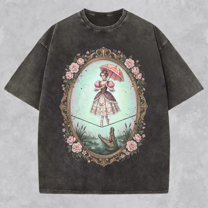 May include: Dark gray t-shirt featuring a vintage-style illustration. The graphic depicts a woman with an umbrella, standing on a tightrope above a swamp with a crocodile. The image is framed by an oval border with pink roses.