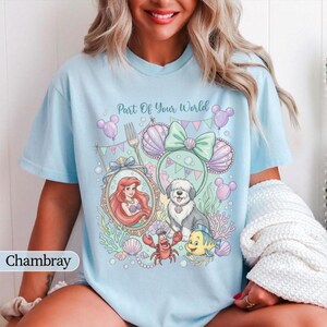 May include: A light blue t-shirt featuring a graphic print with the text Part Of Your World. The design includes a mermaid in an ornate mirror, a dog, a crab, a small fish, seashells, and mouse-ear headbands in pastel shades of pink and green.