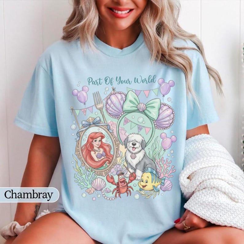 May include: A light blue t-shirt featuring a graphic print with the text Part Of Your World. The design includes a mermaid in an ornate mirror, a dog, a crab, a small fish, seashells, and mouse-ear headbands in pastel shades of pink and green.
