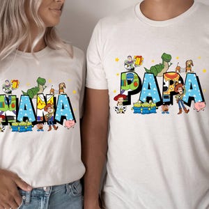 K&ouml;nnte beinhalten: Wei&szlig;e T-Shirts mit den W&ouml;rtern "MAMA" und "PAPA" in gro&szlig;en, farbenfrohen Buchstaben. Die Buchstaben sind mit Zeichentrickfiguren verziert. Die Shirts sind ein passendes Set, perfekt f&uuml;r eine Familie.