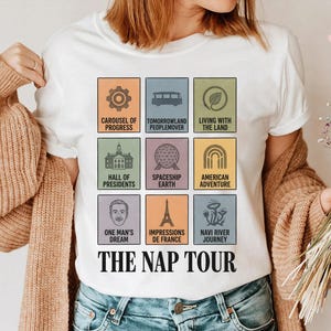 May include: White t-shirt featuring colorful square graphics of theme park attractions, including a carousel, Spaceship Earth, and the Eiffel Tower. The text "THE NAP TOUR" is printed in a bold, black font below the graphics. The shirt is worn by a person.