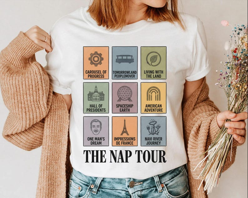May include: White t-shirt featuring colorful square graphics of theme park attractions, including a carousel, Spaceship Earth, and the Eiffel Tower. The text "THE NAP TOUR" is printed in a bold, black font below the graphics. The shirt is worn by a person.