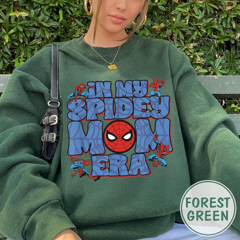 May include: Forest green sweatshirt with the text "IN MY SPIDEY MOM ERA" in blue bubble letters. The design includes Spider-Man graphics and a red Spider-Man mask. The sweatshirt is a comfortable, casual style.