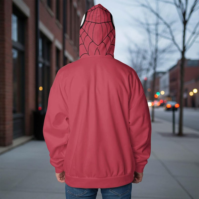 May include: A red hoodie with a Spider-Man mask design on the hood. The hood has a black web pattern and white eye details. The person is wearing blue jeans. The background shows a city street with buildings and trees.