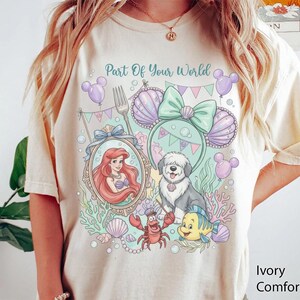 May include: An ivory t-shirt featuring a graphic print with the text Part Of Your World. The design includes a mermaid in a mirror, a dog, a crab, a fish, seashells, and mouse-ear headbands in pastel shades of purple, green, and pink.