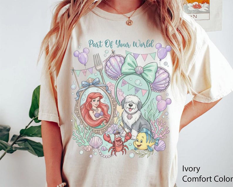 May include: An ivory t-shirt featuring a graphic print with the text Part Of Your World. The design includes a mermaid in a mirror, a dog, a crab, a fish, seashells, and mouse-ear headbands in pastel shades of purple, green, and pink.