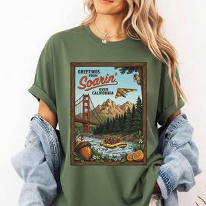 Puede incluir: Camiseta verde oliva con un dise&ntilde;o gr&aacute;fico enmarcado. El gr&aacute;fico muestra el texto "Greetings from Soarin' Over California" y representa el Golden Gate Bridge, monta&ntilde;as, un ala delta y un r&iacute;o con una balsa. Tambi&eacute;n se incluye una bellota y una ardilla de dibujos animados.