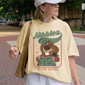 May include: Cream-colored t-shirt featuring a cartoon beaver with the text "Nibbles" and "Gnaw-some". The beaver is wearing a hat and vest, holding a stick. The shirt has a vintage, retro design.