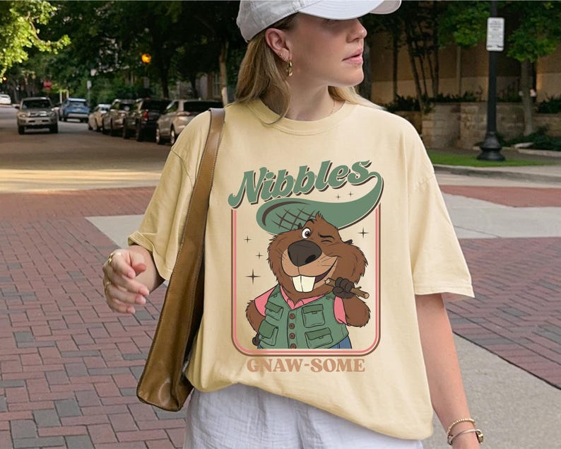 May include: Cream-colored t-shirt featuring a cartoon beaver with the text "Nibbles" and "Gnaw-some". The beaver is wearing a hat and vest, holding a stick. The shirt has a vintage, retro design.