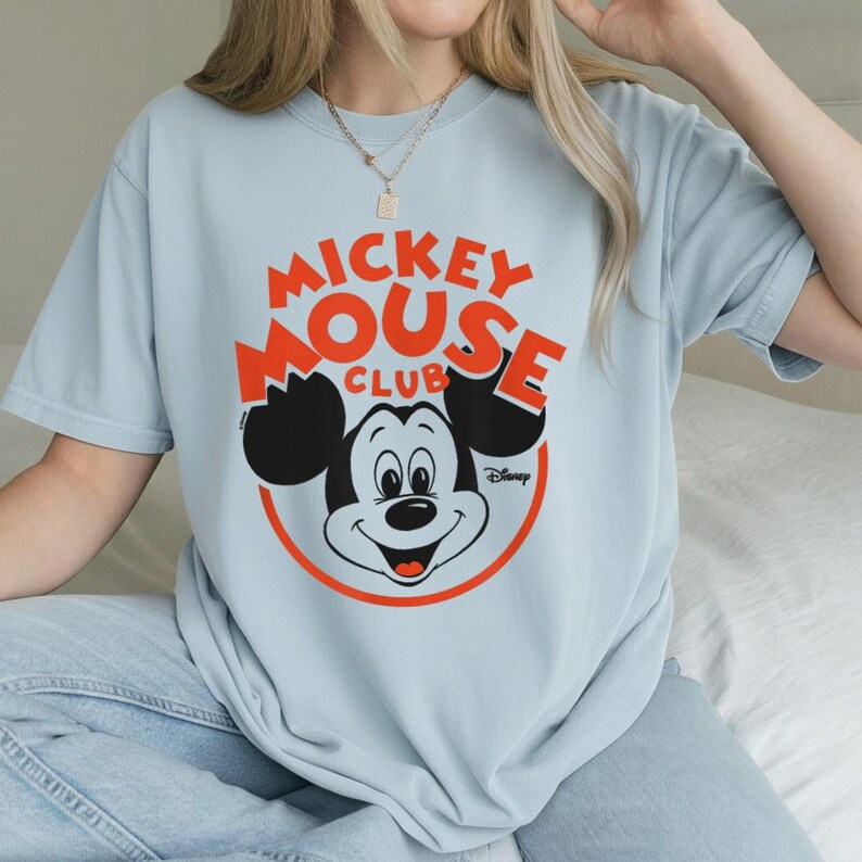 May include: Light blue t-shirt featuring the Mickey Mouse Club logo in orange and black. The design includes Mickey Mouse's face inside a red circle, with the Disney logo below. The shirt is a classic, casual style.