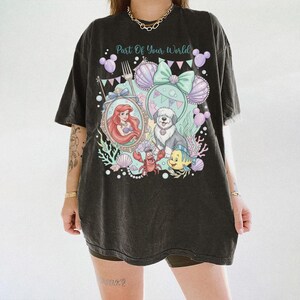 May include: A black oversized t-shirt featuring a graphic print with the text Part Of Your World. The design includes a mermaid, a dog, a crab, a fish, seashells, balloons, and a fork in shades of purple, teal, pink, and white.