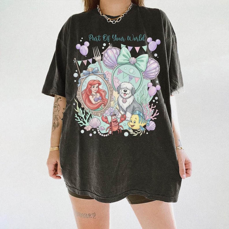 May include: A black oversized t-shirt featuring a graphic print with the text Part Of Your World. The design includes a mermaid, a dog, a crab, a fish, seashells, balloons, and a fork in shades of purple, teal, pink, and white.