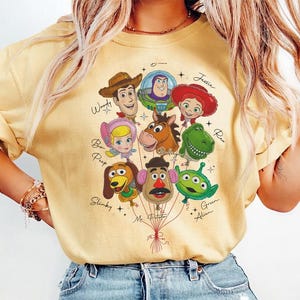 Puede incluir: Camiseta amarilla clara con un gr&aacute;fico de personajes de Toy Story como globos. Los personajes incluyen a Woody, Buzz Lightyear, Jessie, Bo Peep, Slinky, Mr. Potato, Rex y el Alien Verde. La camiseta tiene cuello redondo y mangas cortas.