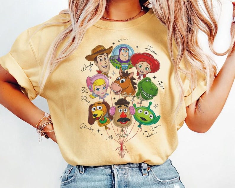 Puede incluir: Camiseta amarilla clara con un gr&aacute;fico de personajes de Toy Story como globos. Los personajes incluyen a Woody, Buzz Lightyear, Jessie, Bo Peep, Slinky, Mr. Potato, Rex y el Alien Verde. La camiseta tiene cuello redondo y mangas cortas.